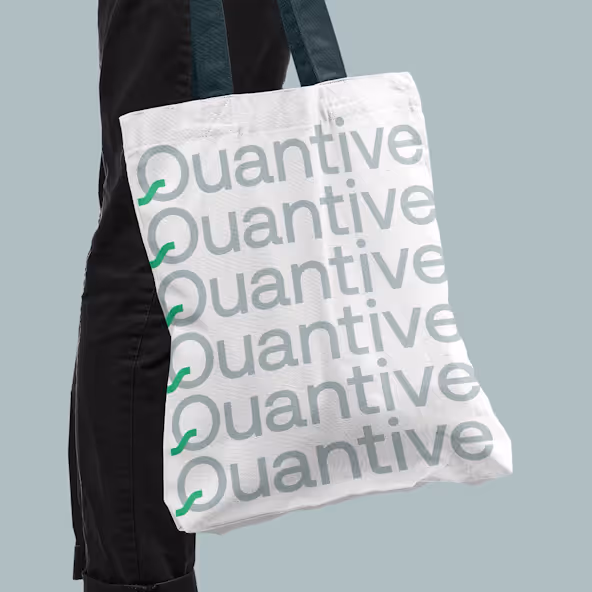 White tote bag with repeated gray text 'Quantive' and green decorative elements held by a person in black pants against a light gray background.