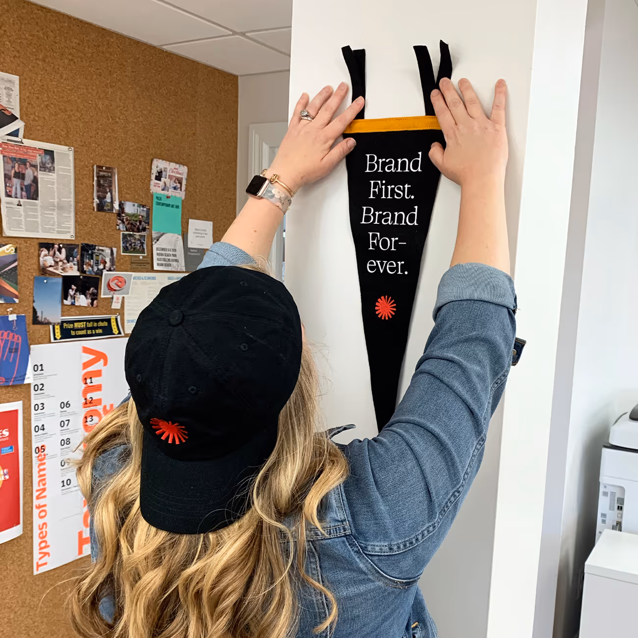 Person with long blonde hair and a black cap hanging a black pennant with the text 'Brand First. Brand Forever.' on a white wall.